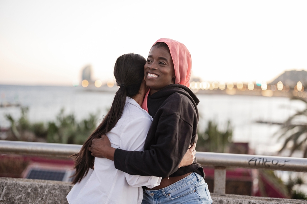 Two Young Women Embracing By The Seaside At Dusk Smiling
